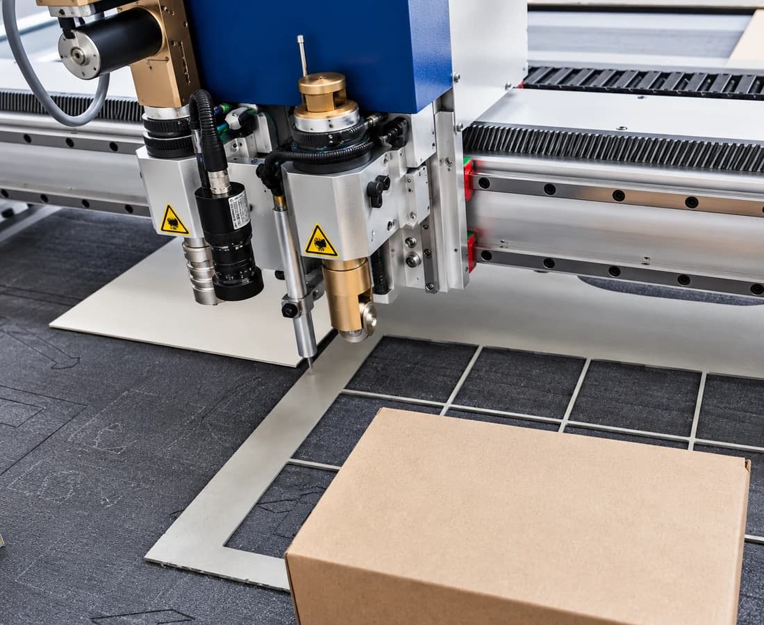 Cabeçote da mesa de corte EK 1625 com a faca oscilante, a câmera CCD, a faca e a roda de vinco sobre o papelão recortado, ao lado da caixa já montada